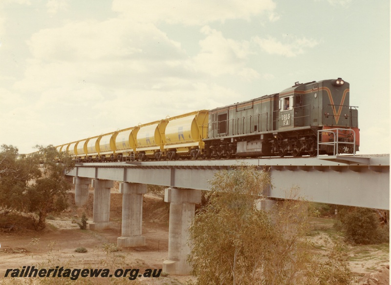 P03875
1 of 2, RA class 1915 diesel locomotive on mineral sands train, crossing the Irwin River bridge, running short hood first, side and front view, in green with red and yellow stripe livery, XE class bogie ilmenite hoppers.DE linne
