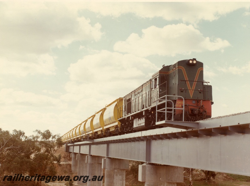 P03876
2 of 2, RA class 1915 diesel locomotive on mineral sands train, crossing the Irwin River bridge, running short hood first, side and front view, in green with red and yellow stripe livery, XE class bogie ilmenite hoppers. DE line
