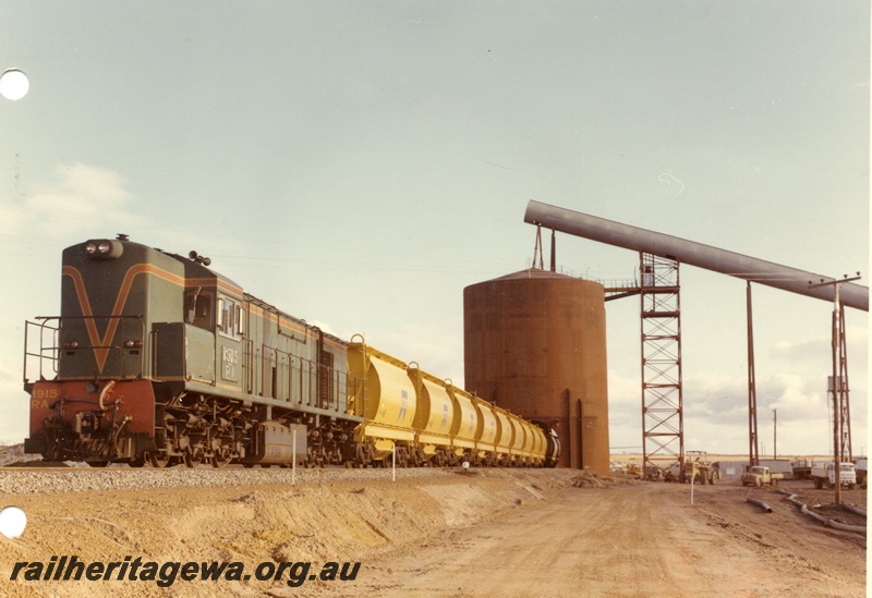 P03880
RA class 1915 diesel locomotive on mineral sands train, running short hood first, front and side view, in green with red and yellow stripe livery, loading XE class bogie ilmenite hoppers, Eneabba. DE line

