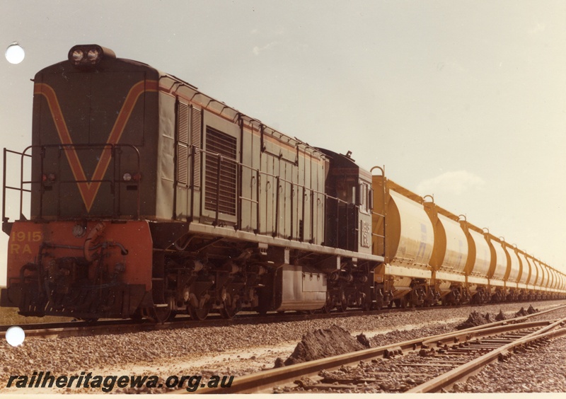 P03881
RA class 1915 diesel locomotive on mineral sands train, running long hood first, front and side view, in green with red and yellow stripe livery, XE class bogie ilmenite hoppers. DE line.
