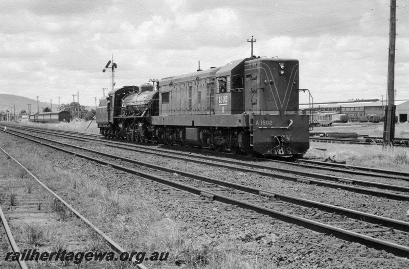 P17152
A class 1502 diesel electric locomotive hauling W class 960 steam locomotive from Midland Workshops to East Perth loco. Note semaphore signal at rear of locos and Workshops buildings at right. Stowed suburban carriages in background. ER line.
