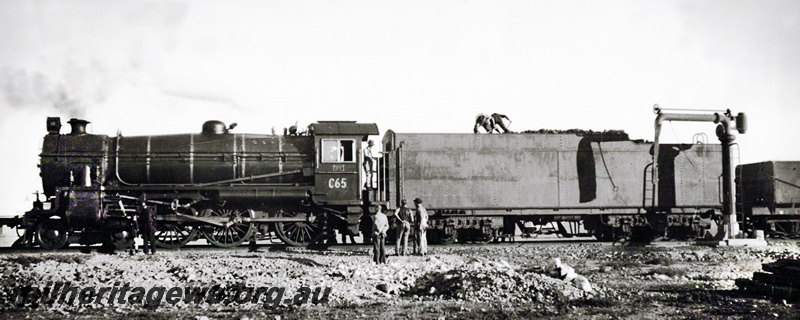 P20910
Commonwealth Railways C class 65 taken water at an unknown location on the Nullarbor.  Side view of the locomotive with the crew on the tender  shovelling coal forward. 
