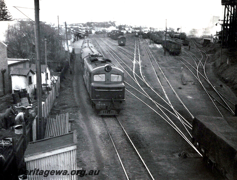 P24035
X class 1008, on passenger train, station building, platform, yard, various wagons, water tower, back fences, Bunbury, SWR line, side and front view from elevated position
